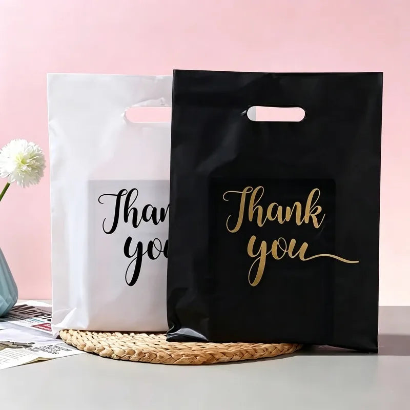 Two black and white gift bags with gold script text saying Thank you are displayed on a light surface.