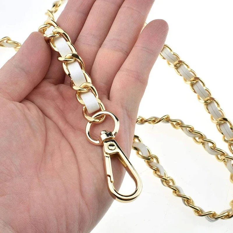 A hand holds a gold-toned metal shoulder strap with white accents, showing its length and texture.