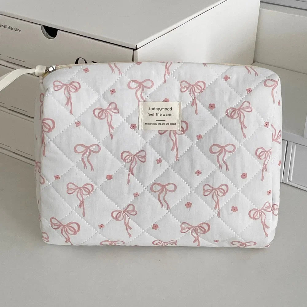 A white quilted cosmetic bag with pink ribbon embroidery, featuring a small tag that reads today mood feel the warm.