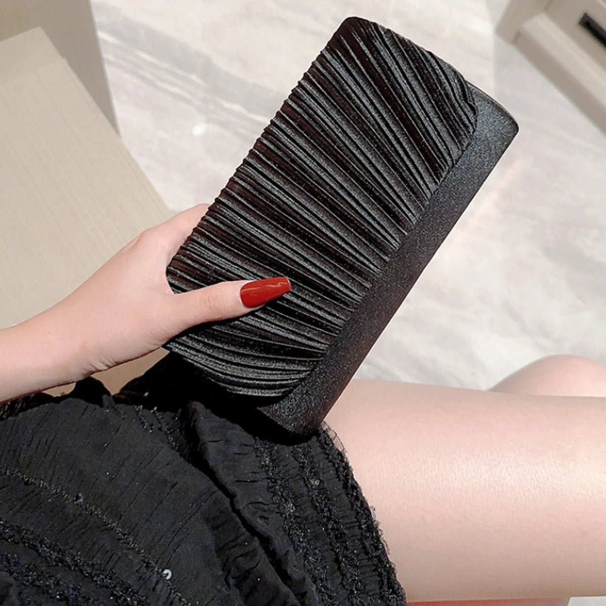 A hand holds a black pleated clutch bag with a textured surface against a light background.