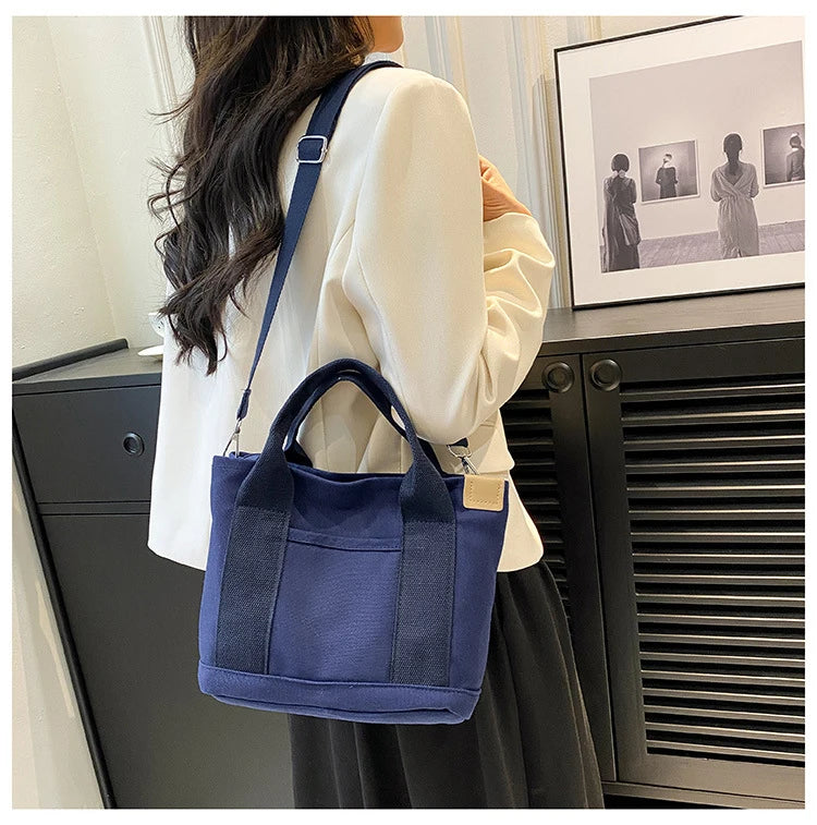 A woman holds a navy blue canvas crossbody bag with an adjustable strap and multiple pockets against a dark background.