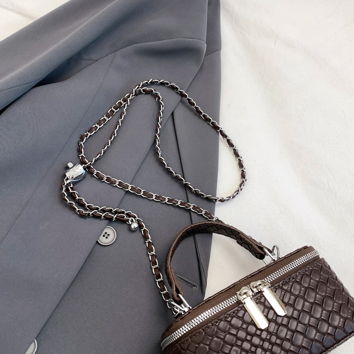 A dark brown woven crossbody bag with a silver chain strap resting on a gray blazer.