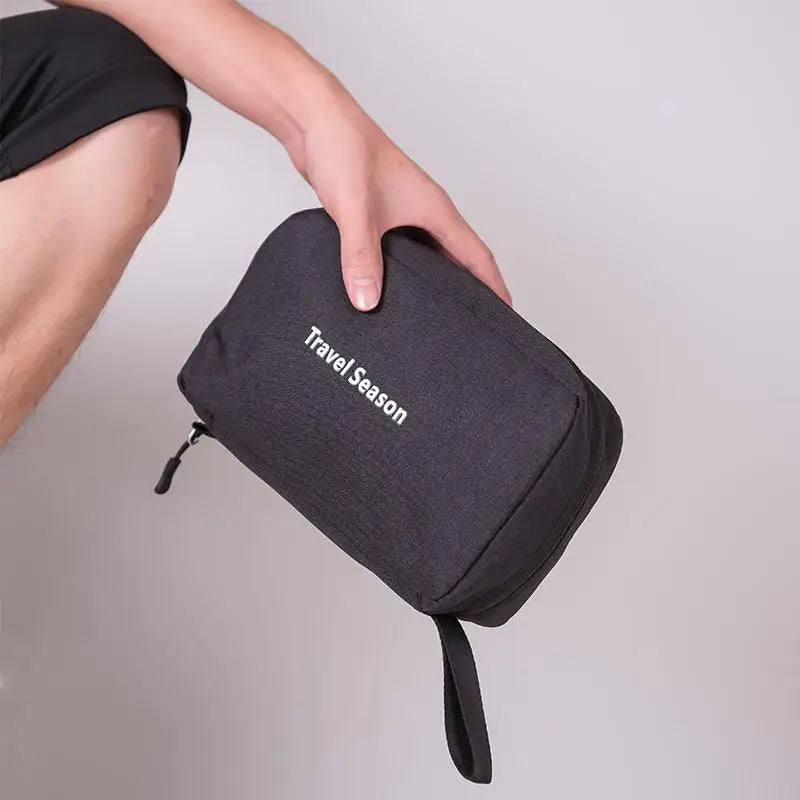 Une main tient un sac de toilette imperméable noir suspendu avec l'inscription Travel Season.
