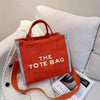 An orange canvas tote bag with white text reading THE TOTE BAG resting on a white knitted surface.