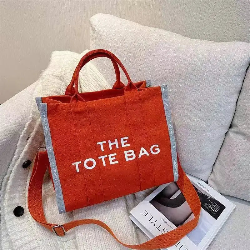 An orange canvas tote bag with white text reading THE TOTE BAG resting on a white knitted surface.