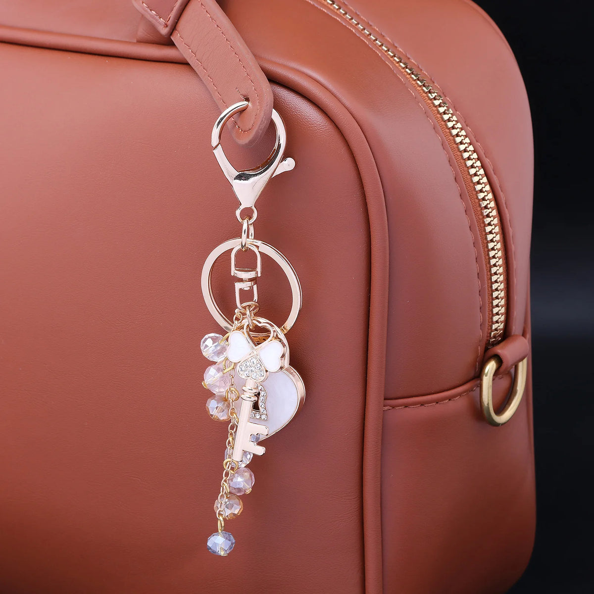 A gold-toned rhinestone bow and heart key chain charm attached to a brown leather handbag.