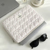 A quilted white laptop sleeve with pink floral embroidery is placed next to a silver laptop and a glass perfume bottle.