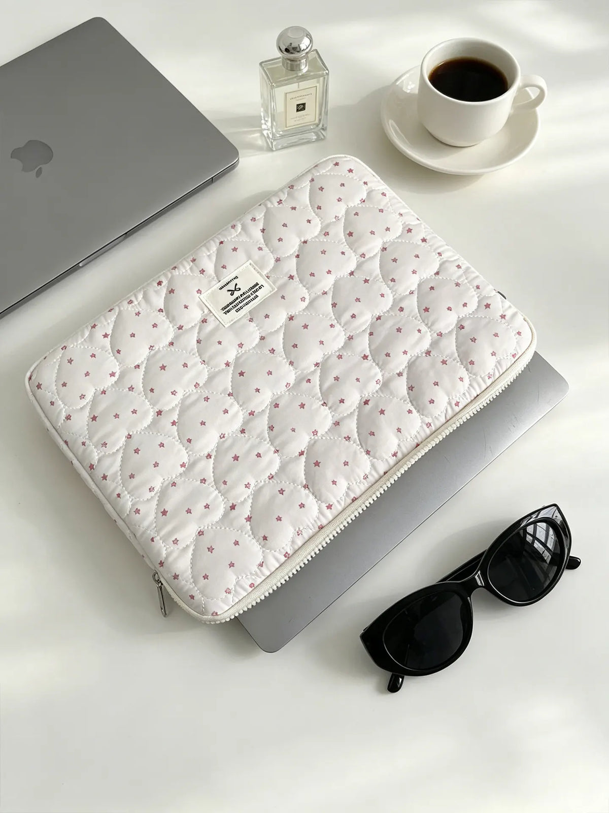 A quilted white laptop sleeve with pink floral embroidery is placed next to a silver laptop and a glass perfume bottle.