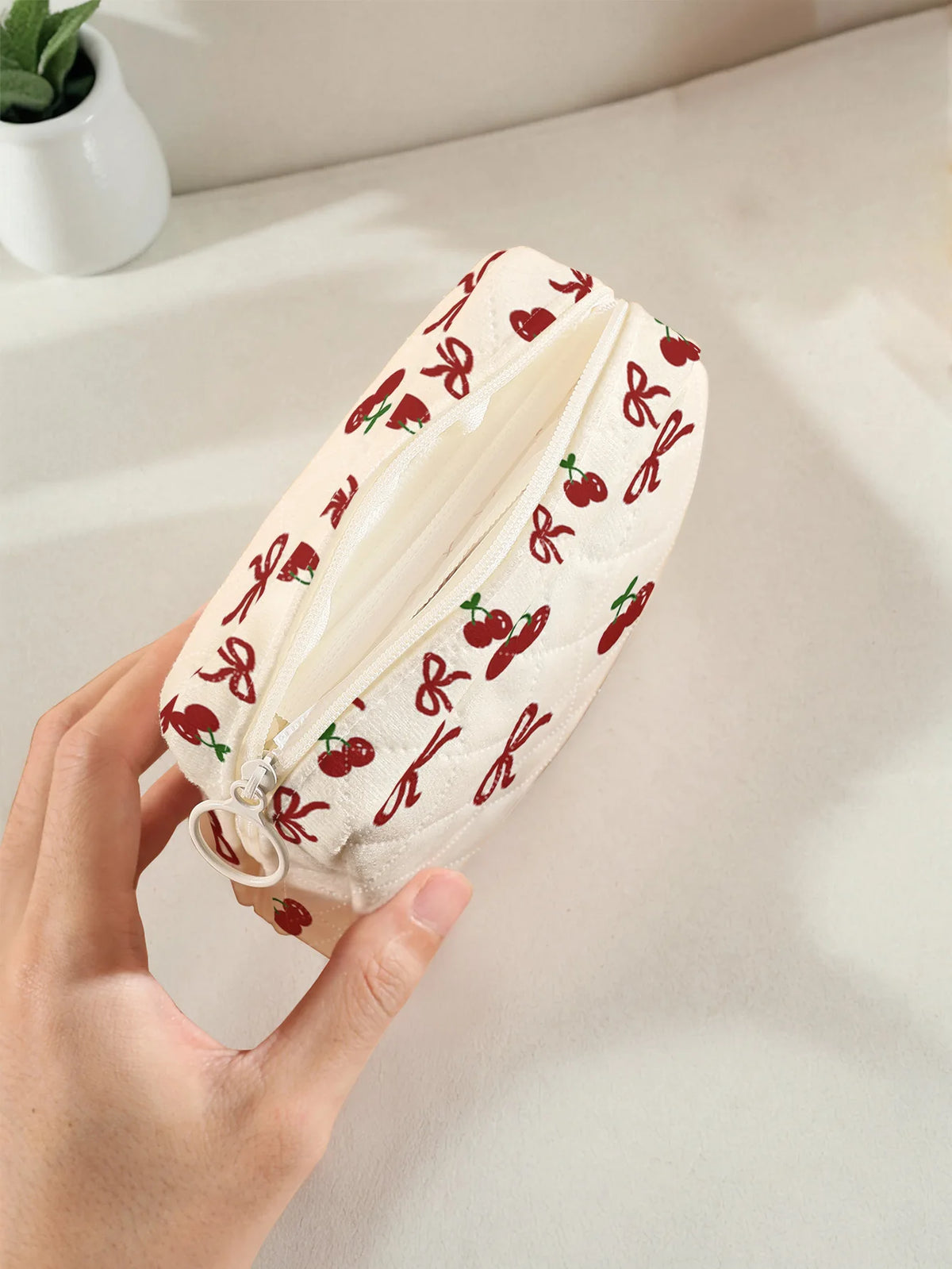 A hand holds a quilted storage pouch with a red cherry and bow print on a light cream fabric.