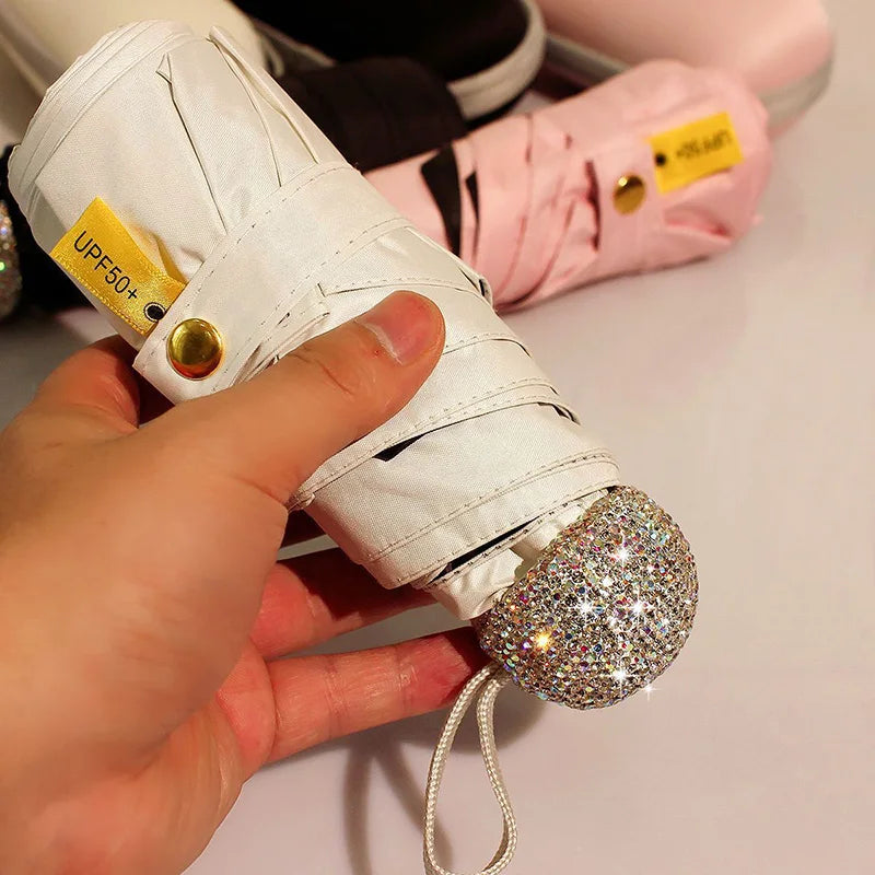 A hand holds a white compact umbrella with gold accents and a rhinestone sphere attached.