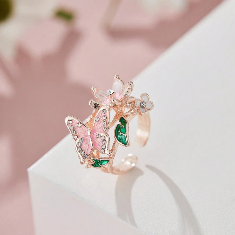 A delicate rose gold ring featuring a pink butterfly and green foliage details.
