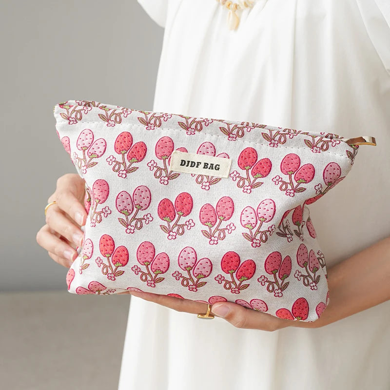 A pink floral canvas makeup bag with a butterfly pattern is held by a person against a white dress.