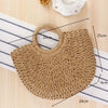 A handwoven straw moon bag with a top handle resting on a striped cloth next to a white cup.