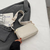 A light beige vegan PU crossbody bag with a woven strap is shown next to a gray blazer on a white surface.