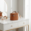 A round, tan travel makeup organizer with a zipper closure sits on a white marble vanity.