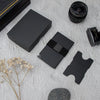 A black RFID blocking metal pop-up card wallet displayed on a white surface with a camera lens and other accessories.
