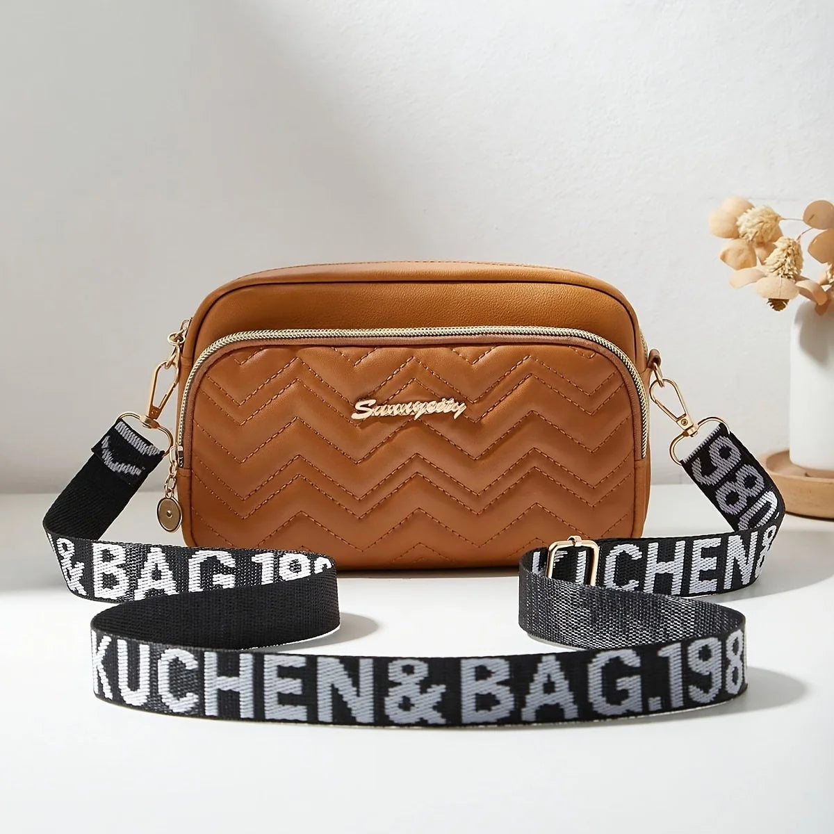 A tan quilted crossbody camera bag with a black and gray letter strap resting on a white surface.