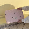 A hand holds a pink quilted slim laptop sleeve for a MacBook or tablet against a bright yellow wall.