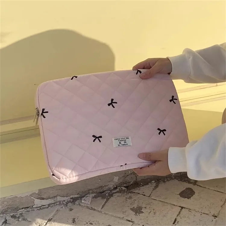 A hand holds a pink quilted slim laptop sleeve for a MacBook or tablet against a bright yellow wall.