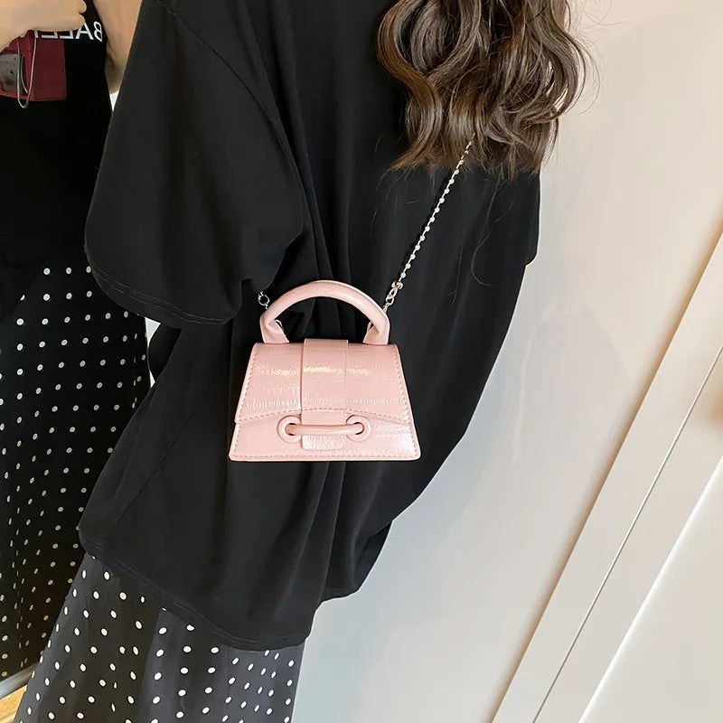A pink leather square crossbody bag with a silver chain strap is shown being held by a person against a white background.