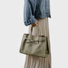 A woman wearing a denim jacket and light pleated skirt holds a beige genuine leather shoulder tote bag.
