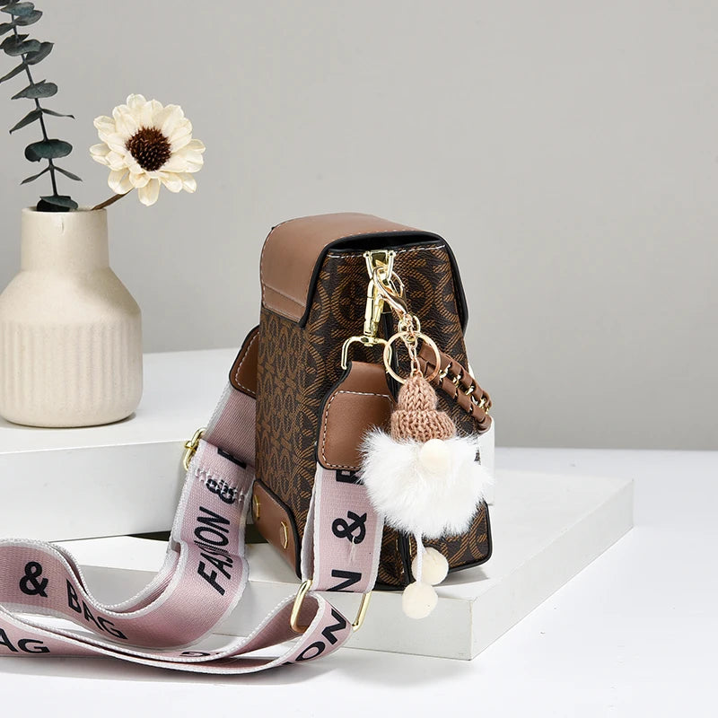 Sac bandoulière en similicuir marron avec quincaillerie dorée et un pompon blanc moelleux sur une surface blanche.