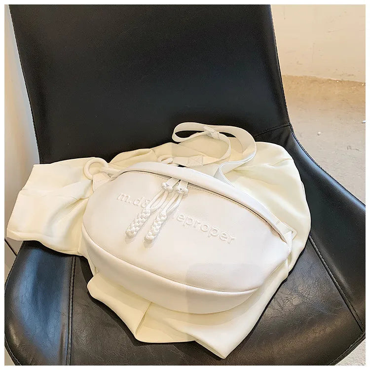 A cream-colored PU leather crossbody saddle bag with a white chain strap resting on a black leather chair.