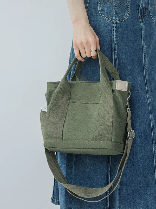 A woman holds a olive green canvas crossbody bag with a tan strap against a light gray background.