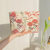 A hand holds a floral canvas makeup bag featuring red and green flowers against a white background.