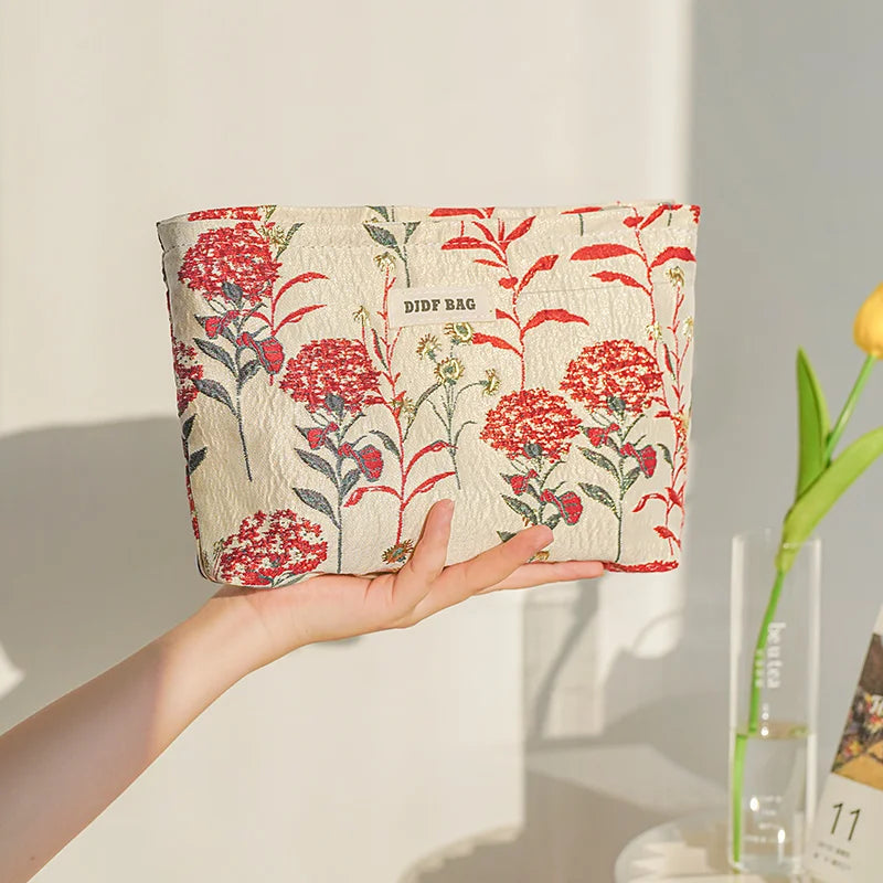 A hand holds a floral canvas makeup bag featuring red and green flowers against a white background.