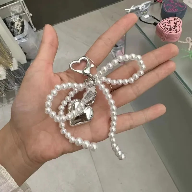 A hand holds a pearl bowknot keychain featuring a heart charm and a small silver charm.