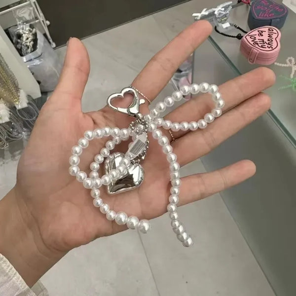 A hand holds a pearl bowknot keychain featuring a heart charm and a small silver charm.