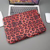 Leopardmuster-Laptophülle in leuchtendem Rot auf grauem Untergrund neben einem geöffneten Laptop.