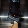A close-up shot of a black genuine leather square shoulder crossbody bag with gold hardware, held by a person wearing dark cl