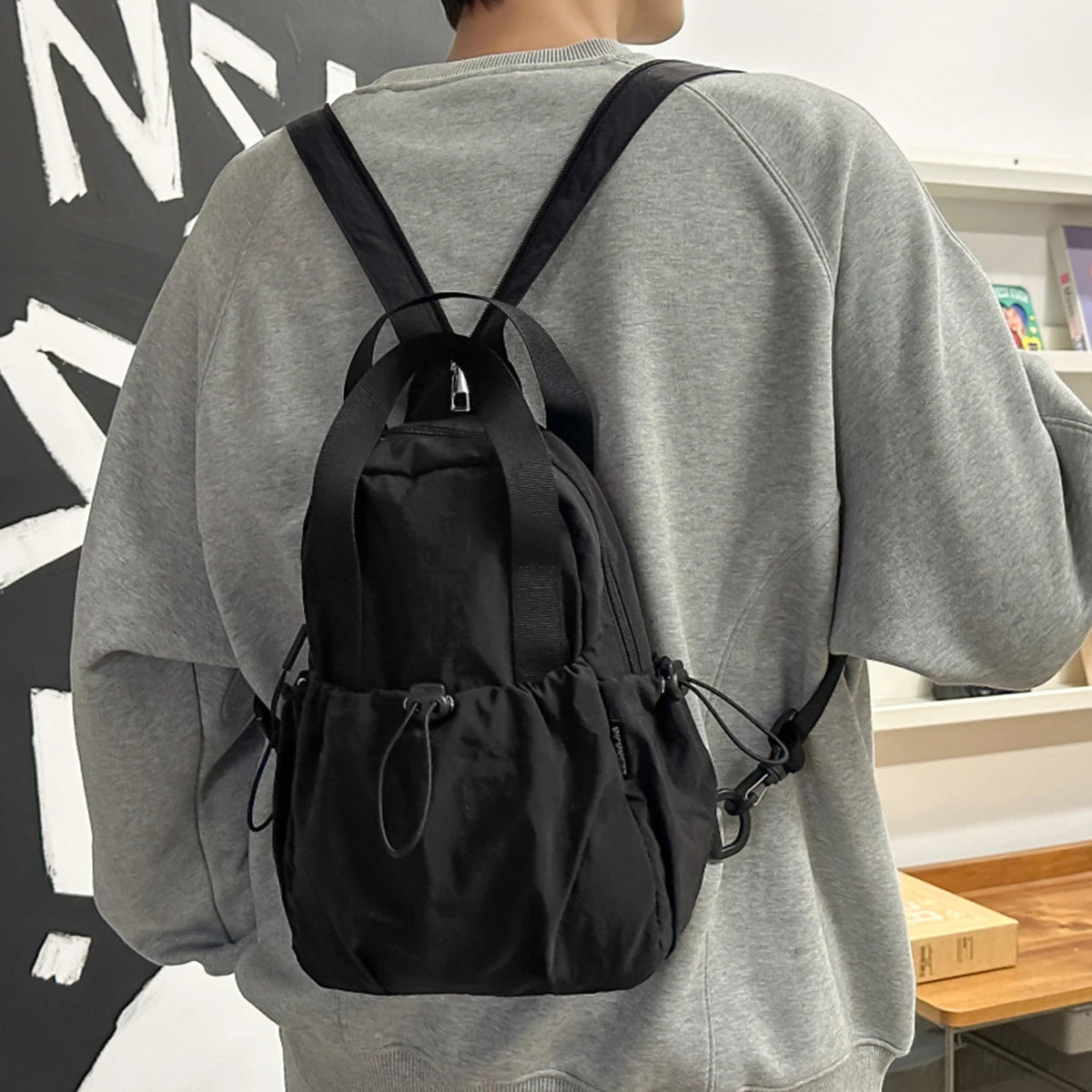 A person wearing a gray sweatshirt is shown from the back, carrying a black casual backpack with shoulder straps.