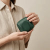 A person holds a dark green compact coin purse with a snap closure against a light background.