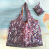 A purple and pink patterned reusable shopping bag is displayed against a light blue background with water and a starfish.