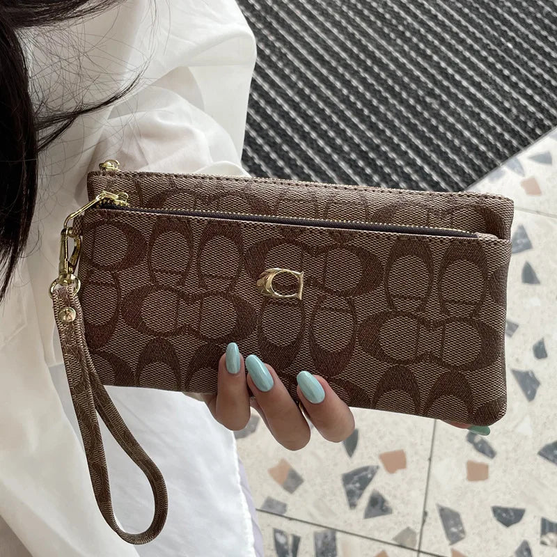A brown patterned zippered wallet is held by a person against a light background.