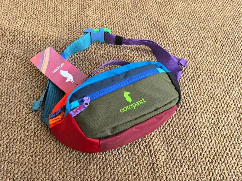 Durable Outdoor Sports Crossbodybag in purple and green with a pink tag on a brown textured surface.