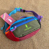 Durable Outdoor Sports Crossbodybag in purple and green with a pink tag on a brown textured surface.