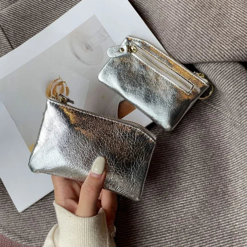 A silver metallic leather compact coin purse and key holder is shown being held against a gray fabric background.