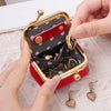 Una mano abre una caja de almacenamiento de joyas de terciopelo con borde dorado y forro rojo para revelar anillos y pendient