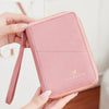 A hand holds a pink RFID-blocking passport wallet with a zipper closure, featuring a gold foil logo.