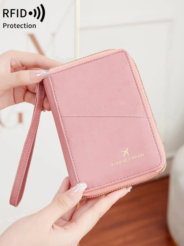 A hand holds a pink RFID-blocking passport wallet with a zipper closure, featuring a gold foil logo.