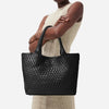 A woman holds a black woven leather tote bag with a handle against a light-colored knit dress.