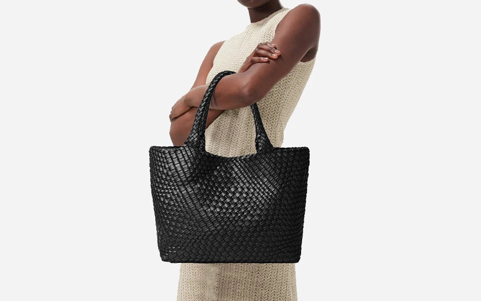 A woman holds a black woven leather tote bag with a handle against a light-colored knit dress.