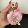 A hand holds a custom initial letter keychain featuring a pink fluffy charm and a sparkly resin pendant.