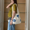 A woman wearing a olive green shirt and denim overalls holds a white knitted tote bag with blue floral embroidery.