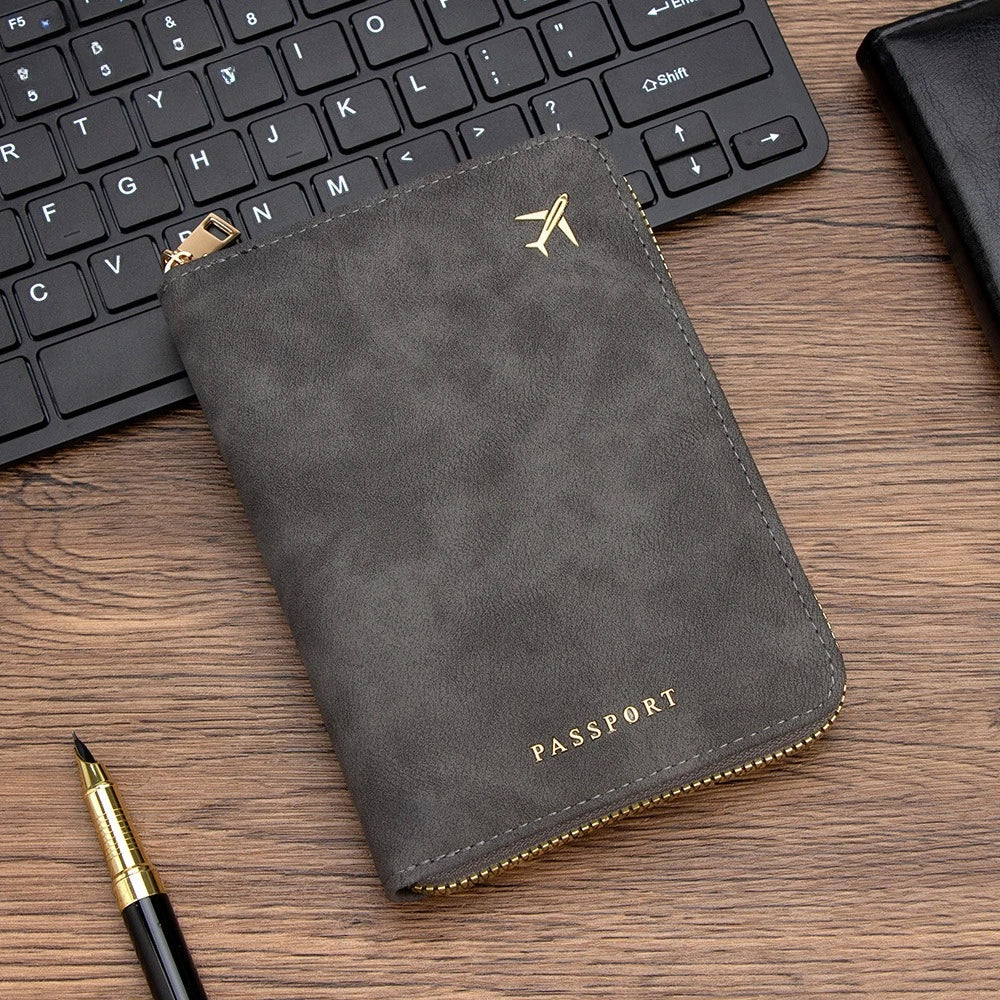 A dark gray slim passport wallet with gold accents rests on a wooden surface next to a laptop keyboard and a pen.