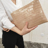A woman holds a beige alligator print clutch bag against a light background.